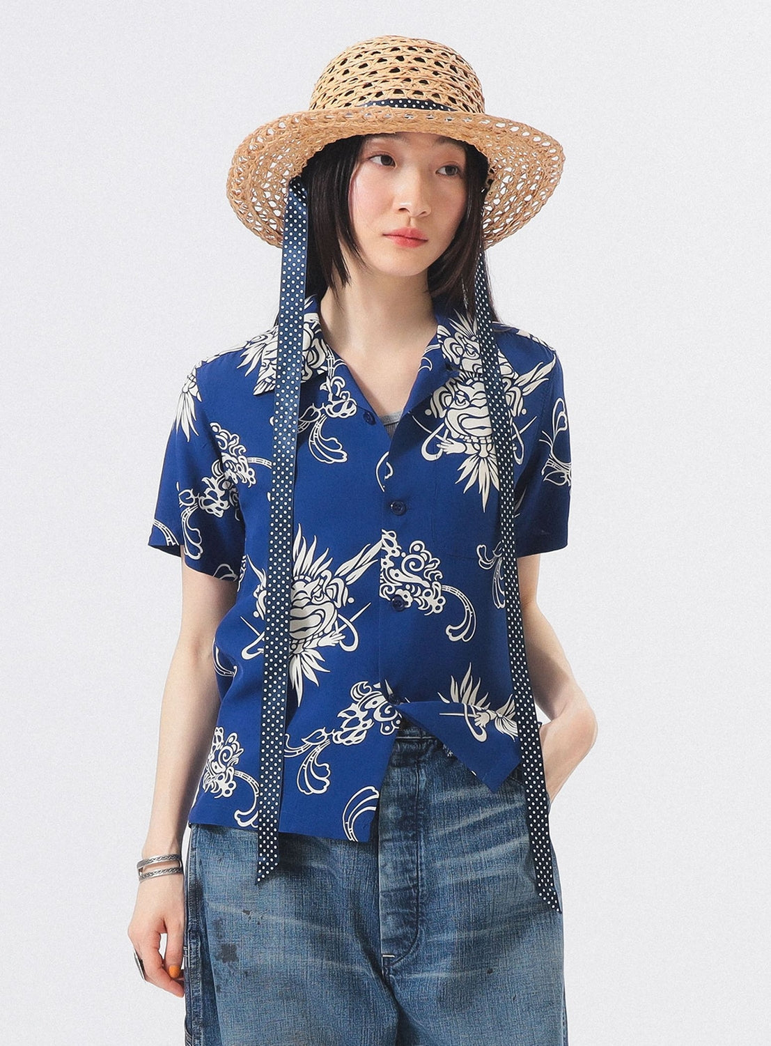A woman models the [Exclusive] SUN SURF x BEAMS BOY Open Collar Shirt by SUN SURF— a blue floral vintage-inspired look—paired with wide-leg denim jeans and a straw hat, standing against a plain white background.