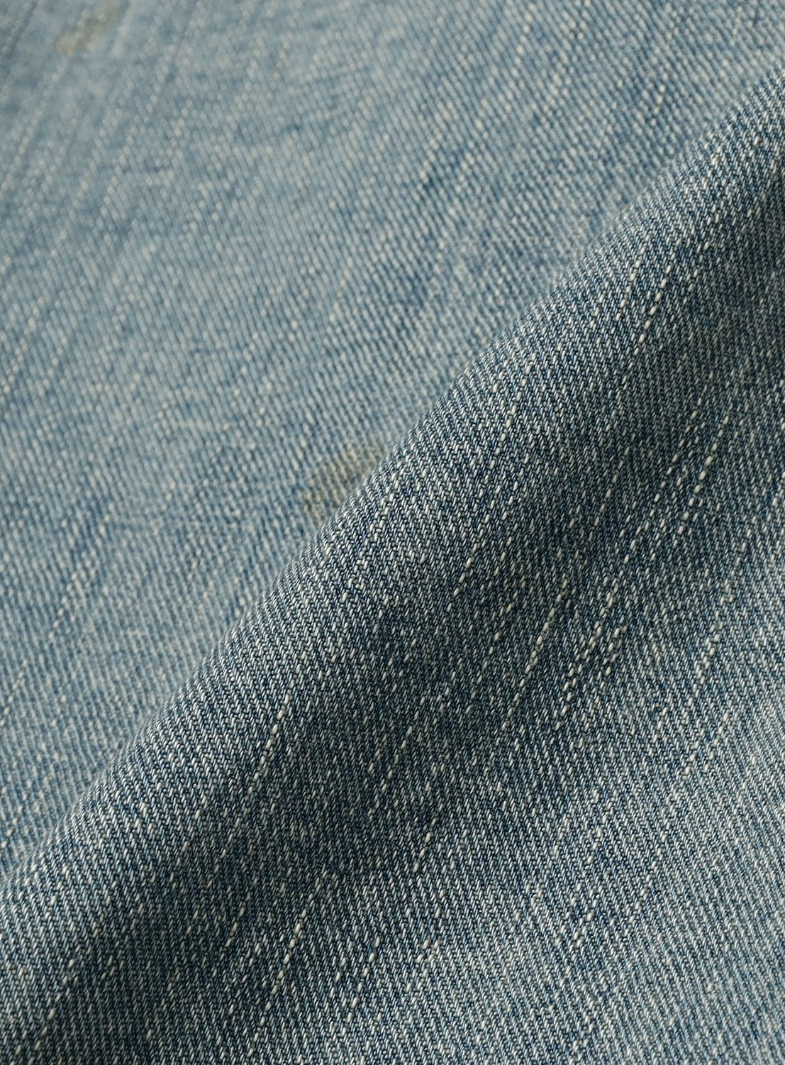 Close-up of SUGARHILL's Faded Tyrol Denim Jacket shows light blue denim with visible weave and vintage wash. Diagonal lines, texture, and a gentle fold add depth and a classic touch to the surface.