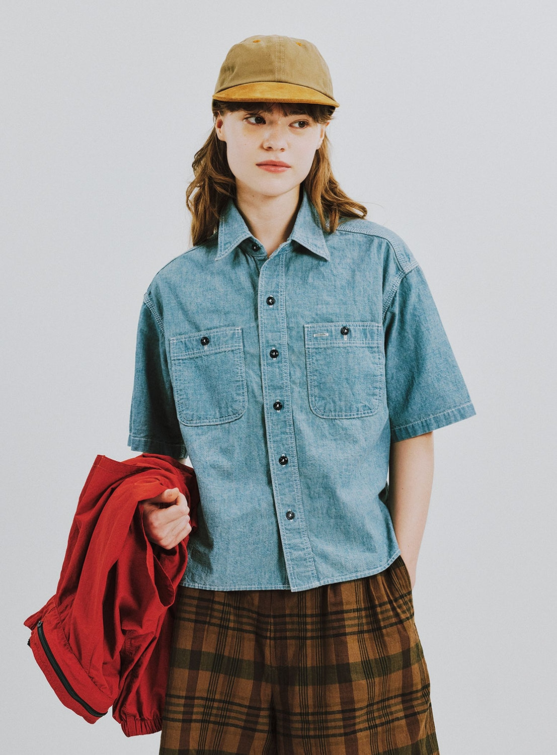 A person wears a tan cap, BEAMS BOY Chambray Short-Sleeve Work Shirt, and brown plaid skirt, holding a red jacket against a plain white background.