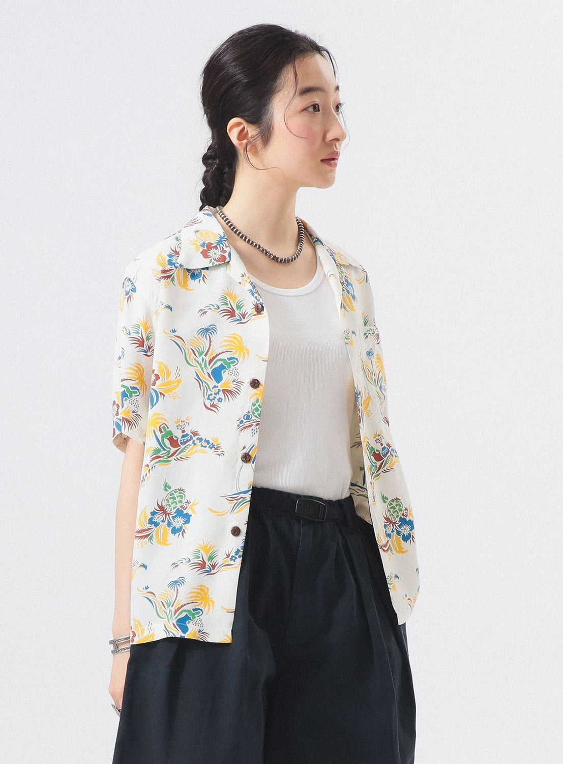 A woman with dark braided hair models the [Exclusive] SUN SURF x BEAMS BOY Open Collar Shirt by SUN SURF over a white top, paired with high-waisted black pants and a necklace, set against a plain light background.