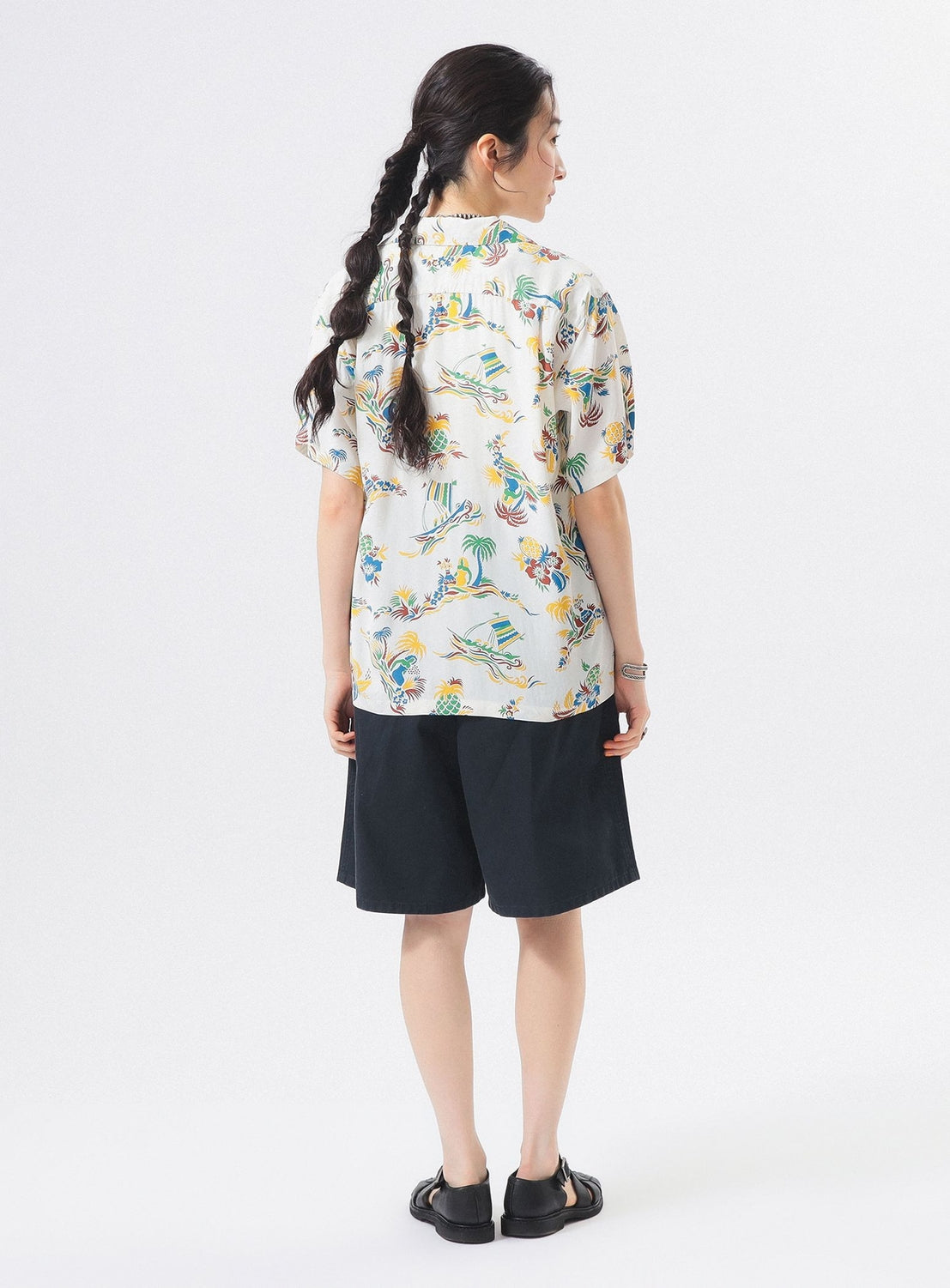 A person with long braided hair stands facing away, wearing the [Exclusive] SUN SURF x BEAMS BOY Open Collar Shirt by SUN SURF, black shorts, and sandals—embodying BEAMS BOY’s laid-back style against a plain white background.