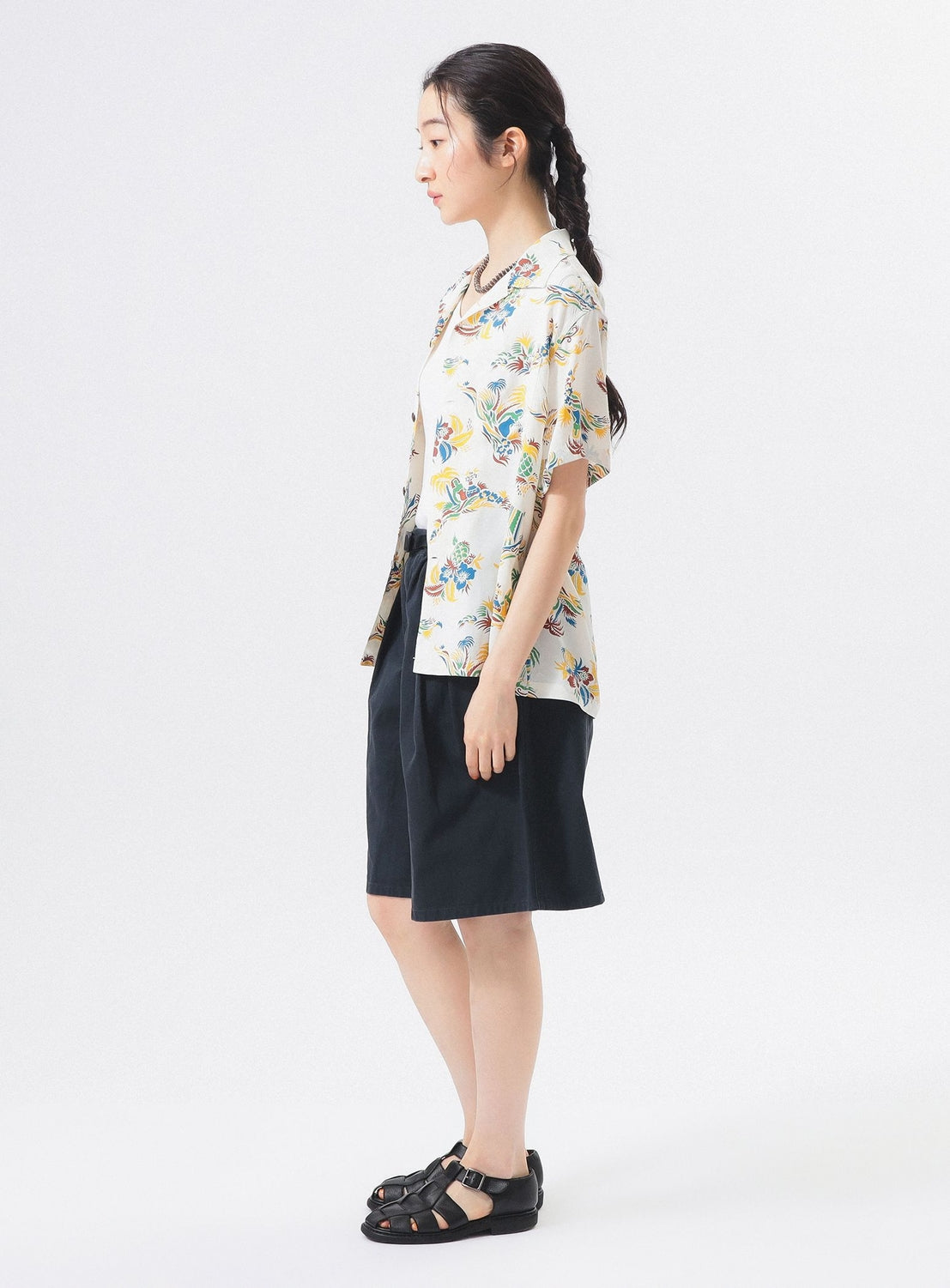 A woman with braided hair stands in profile against a plain background, wearing the [Exclusive] SUN SURF x BEAMS BOY Open Collar Shirt by SUN SURF, knee-length black shorts, black sandals, and a crossbody bag.