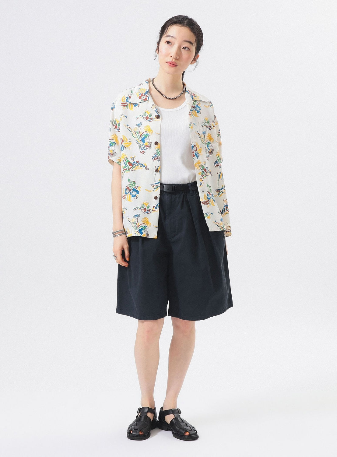 A person stands against a white background wearing the [Exclusive] SUN SURF x BEAMS BOY Open Collar Shirt by SUN SURF over a white top, with dark knee-length shorts, black sandals, and a necklace for a relaxed Hawaiian style.
