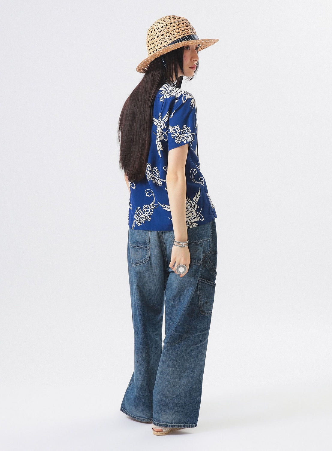 A person stands facing away, wearing the [Exclusive] SUN SURF x BEAMS BOY Open Collar Shirt by SUN SURF, wide-leg denim jeans, a straw hat, and sandals. Their long dark hair flows down their back against a plain white background.