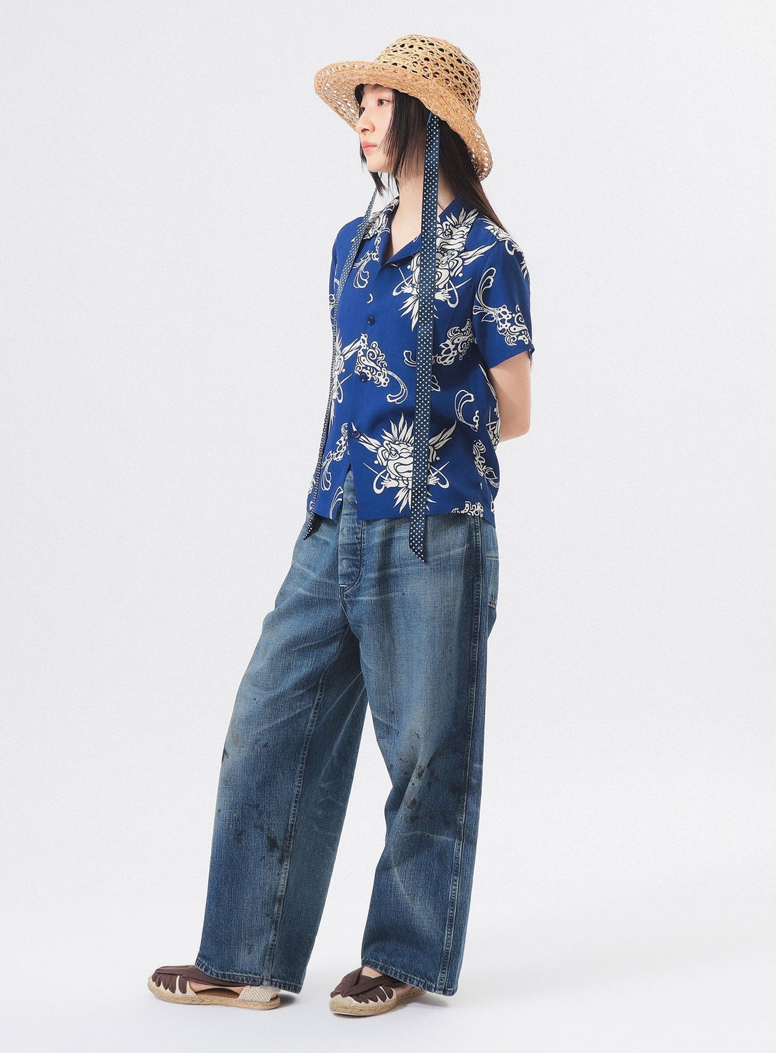 A person wears the [Exclusive] SUN SURF x BEAMS BOY Open Collar Shirt by SUN SURF, loose blue jeans, brown shoes, and a straw hat with long ribbons, standing against a white background and looking to the side.