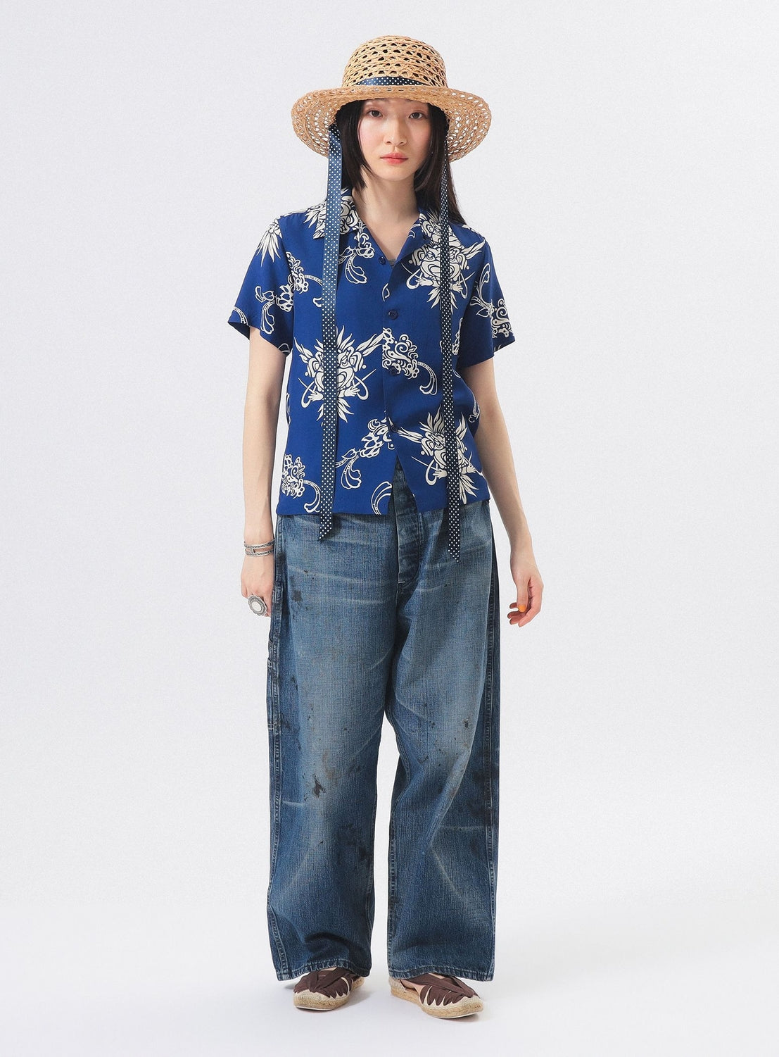 A person in a wide-brimmed straw hat with long black ties, the [Exclusive] SUN SURF x BEAMS BOY Open Collar Shirt by SUN SURF, baggy faded jeans, and brown shoes stands against a plain white background.