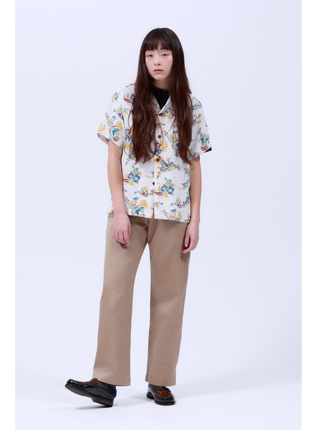 A person with long brown hair stands against a plain background, wearing a white [Exclusive] SUN SURF x BEAMS BOY Open Collar Shirt by SUN SURF in Hawaiian style with a colorful floral print, paired with beige trousers and black shoes.