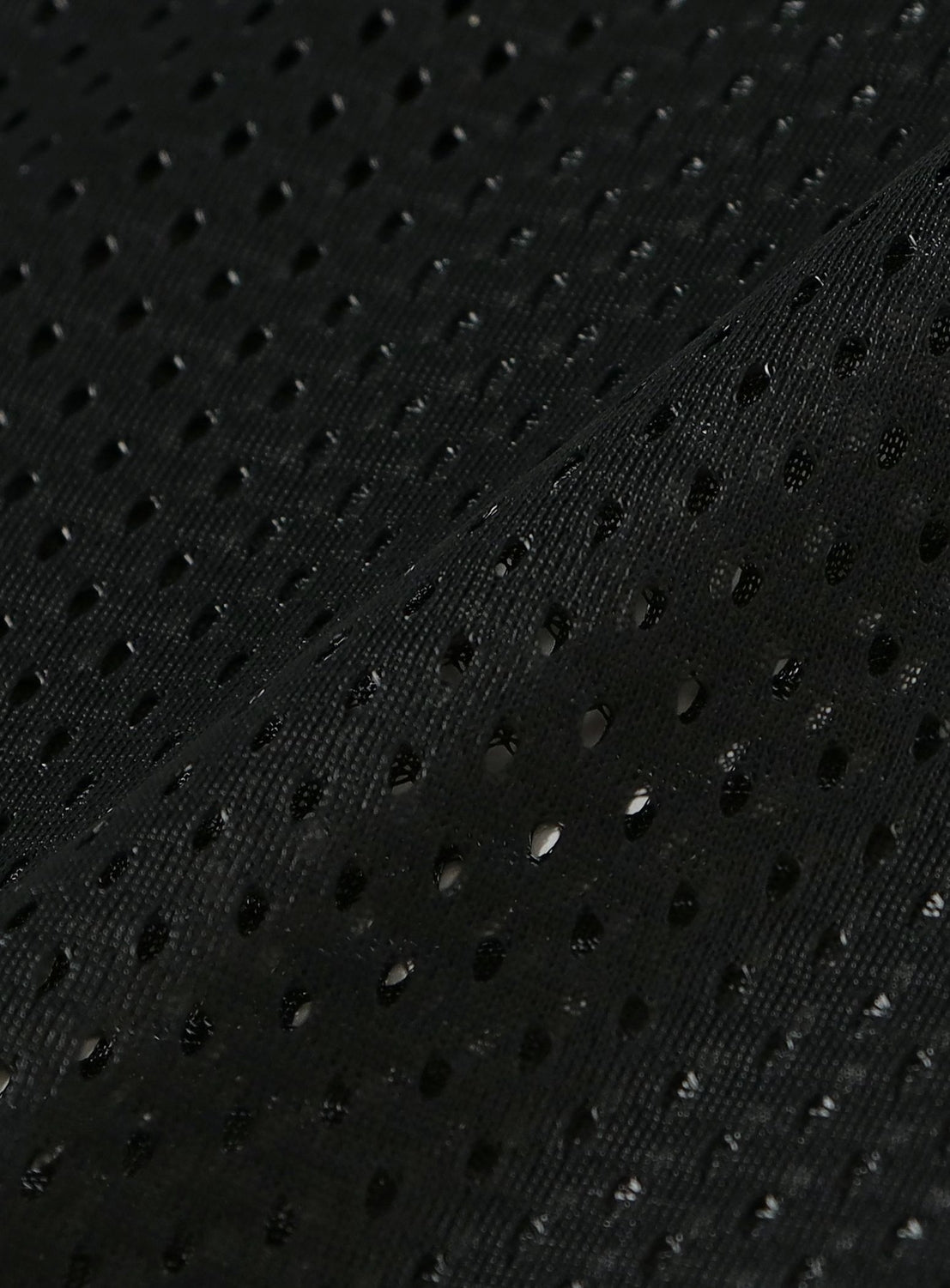 Close-up of black fabric from the [Exclusive] Needles 50th Baseball Shirt by NEEDLES, featuring evenly spaced small holes that create a breathable mesh texture typical of streetwear baseball shirts. The material overlaps diagonally, forming a subtle line across the image.