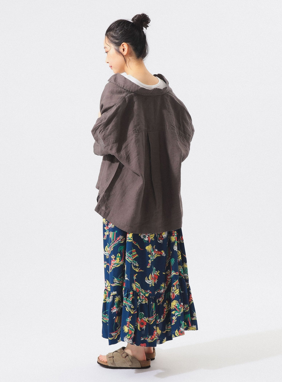 A woman with her hair in a bun stands sideways, looking down, wearing the [Exclusive] SUN SURF Sleeveless Cami Dress by SUN SURF, featuring colorful Hawaiian floral patterns, with a loose brown jacket and brown sandals against a white background.