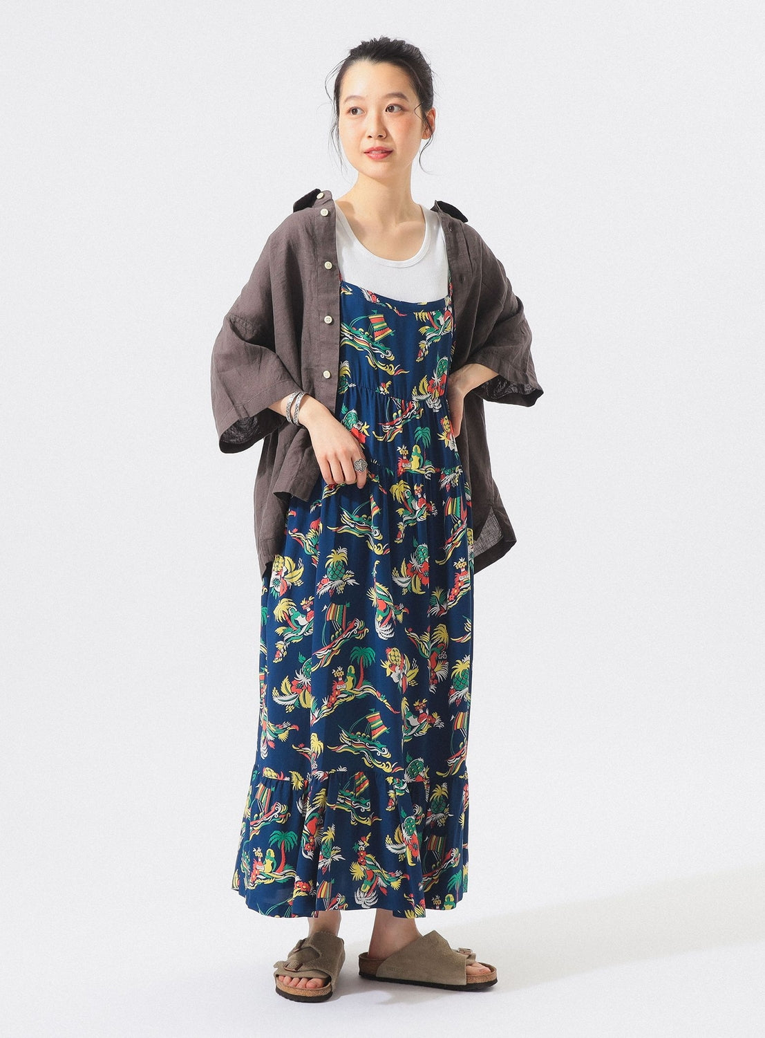 A person stands smiling in the [Exclusive] SUN SURF Sleeveless Cami Dress by SUN SURF—a blue floral maxi dress styled as resort wear—paired with a white top, loose brown button-up shirt, and beige sandals against a plain white background.