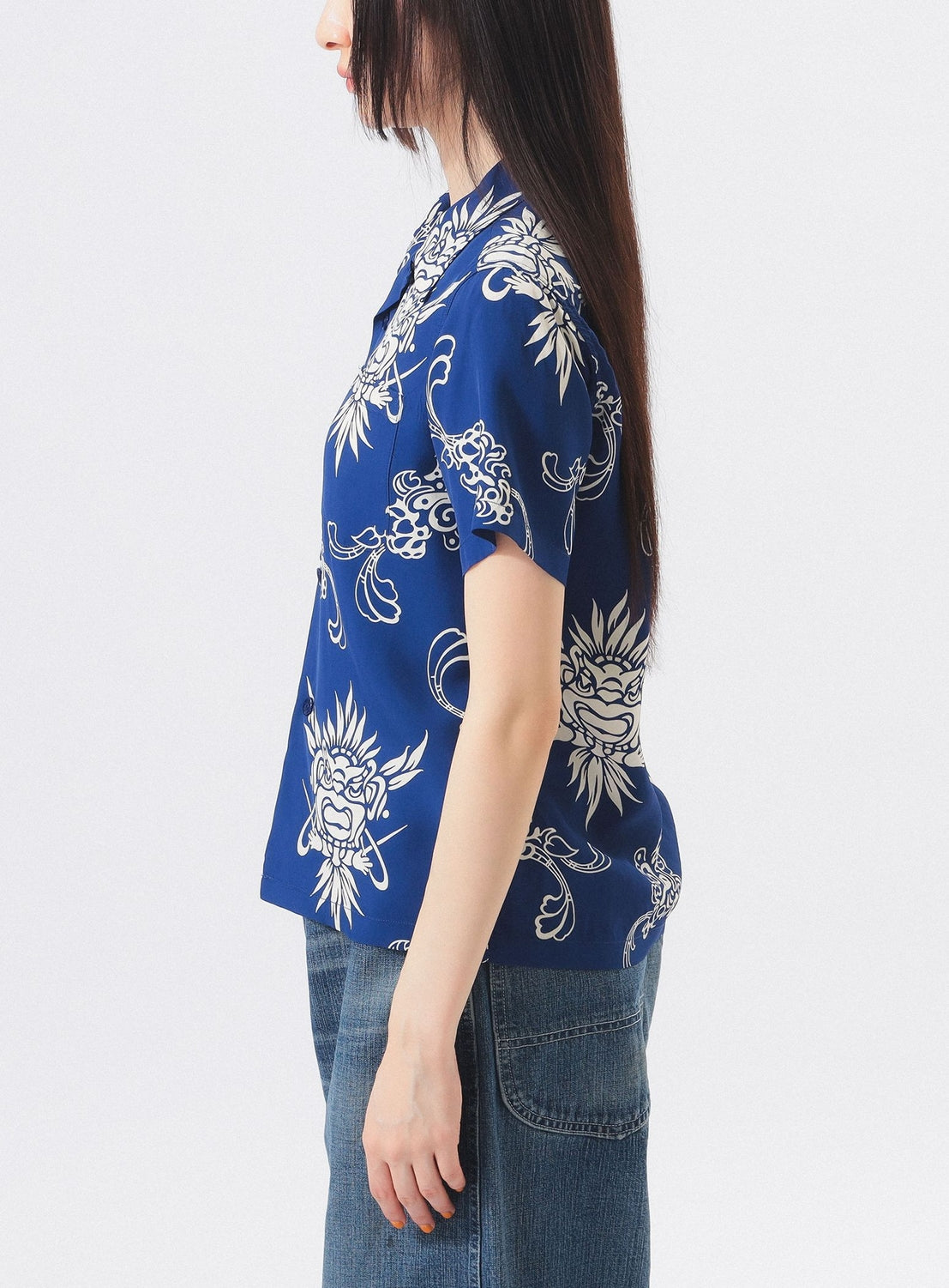 A woman stands in profile wearing the [Exclusive] SUN SURF x BEAMS BOY Open Collar Shirt from SUN SURF—a blue, short-sleeved aloha shirt with a white abstract pattern—paired with blue jeans against a plain light background.