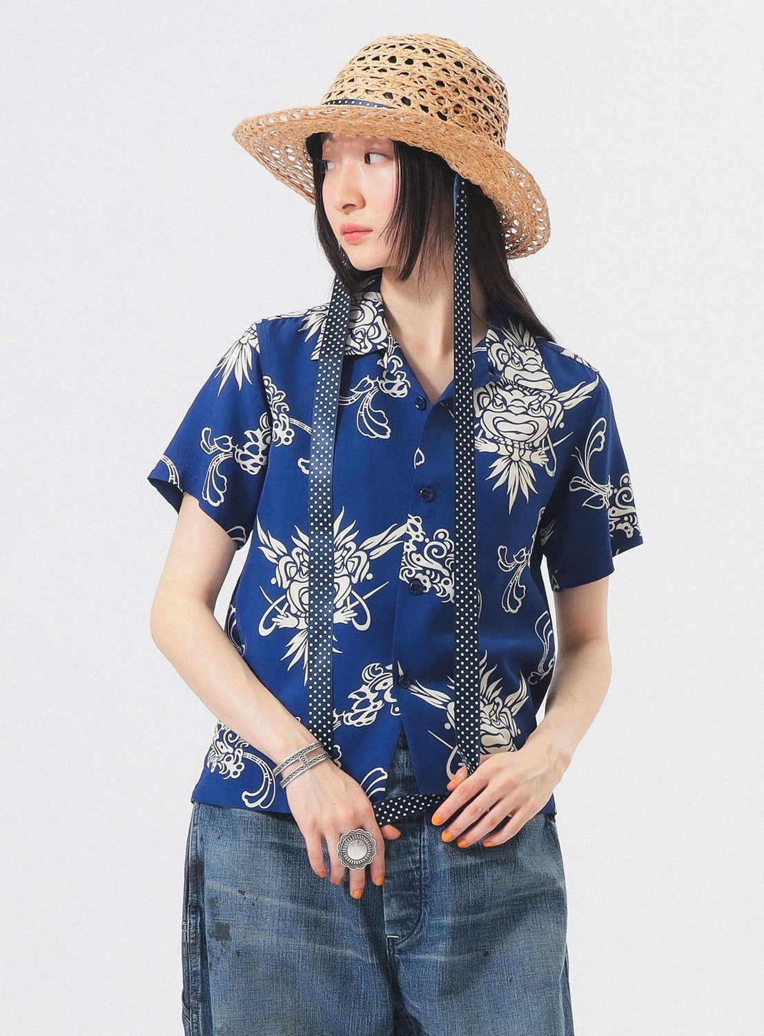A person wears the SUN SURF x BEAMS BOY Open Collar Shirt by SUN SURF, featuring blue vintage style with white floral patterns, paired with a wide-brimmed straw hat and faded jeans against a plain light background.