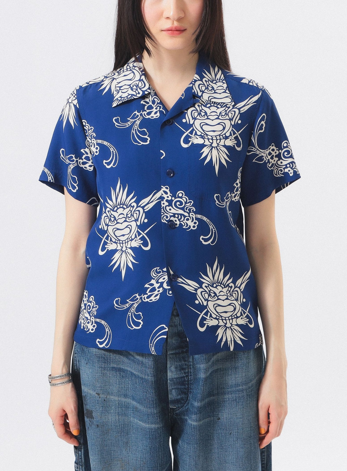 A person wears the [Exclusive] SUN SURF x BEAMS BOY Open Collar Shirt by SUN SURF—a blue, short-sleeve vintage aloha shirt with bold white graphics—and blue jeans, standing against a plain white background with their face partially out of frame.