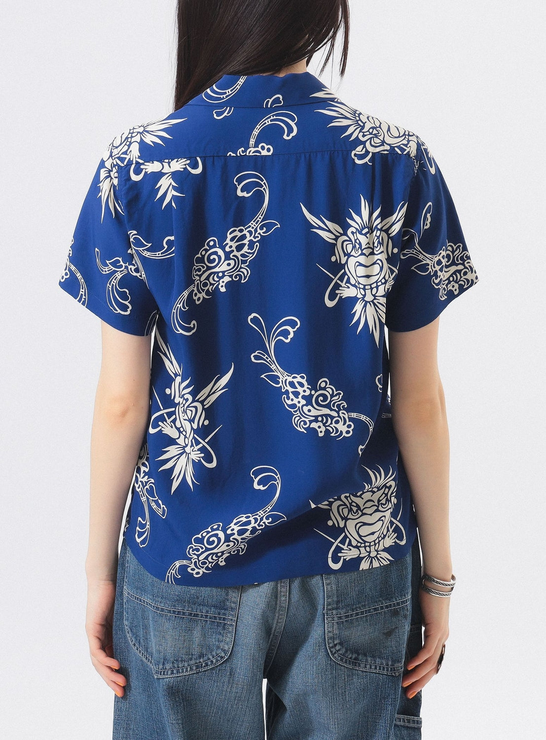 A person with long dark hair faces away from the camera, wearing the [Exclusive] SUN SURF x BEAMS BOY Open Collar Shirt by SUN SURF, styled with blue denim jeans and bracelets for a relaxed Hawaiian-inspired look.