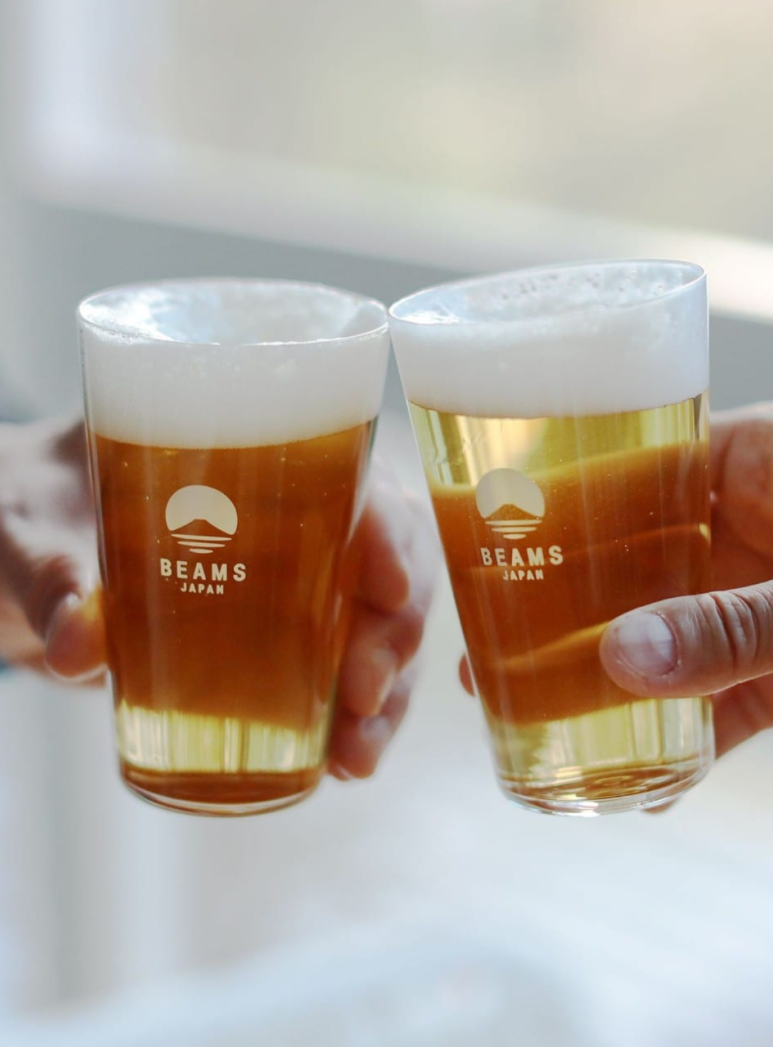 Two glasses of beer with 'BEAMS JAPAN' logo held by hands against a blurred background