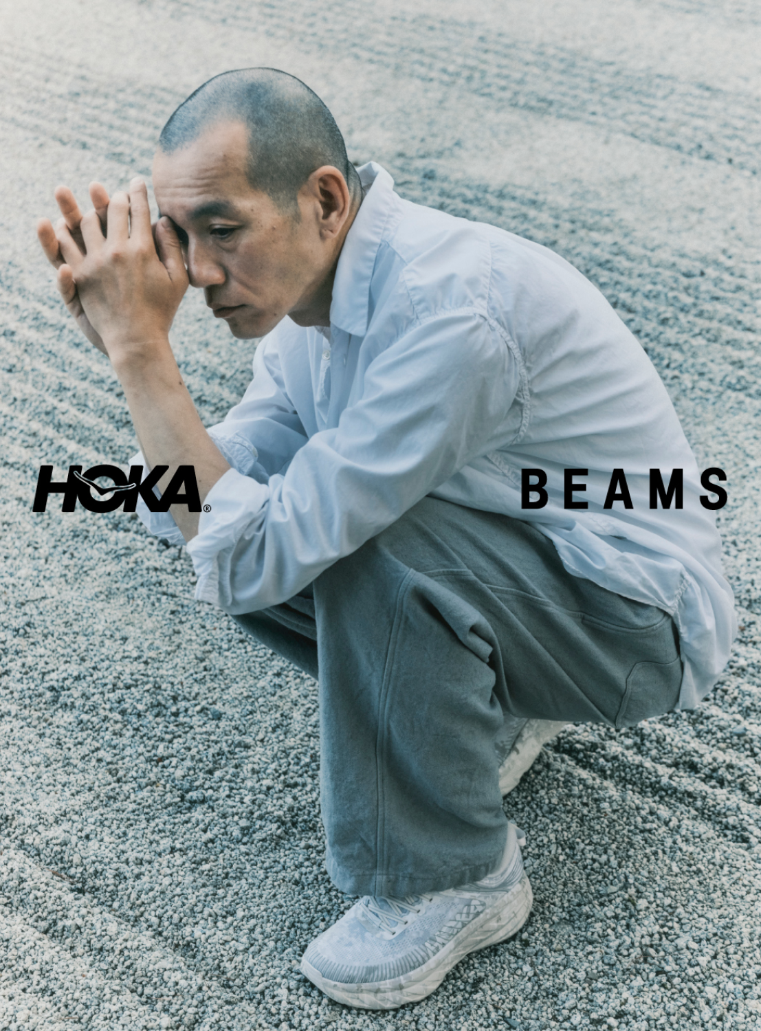 A man in a white shirt and gray pants squats on gravel with his hands touching his face, wearing the Hoka U BONDI 7 BEAMS sneakers, known for their engineered mesh upper and lightweight cushioning. The Hoka and BEAMS logos are displayed over the image.