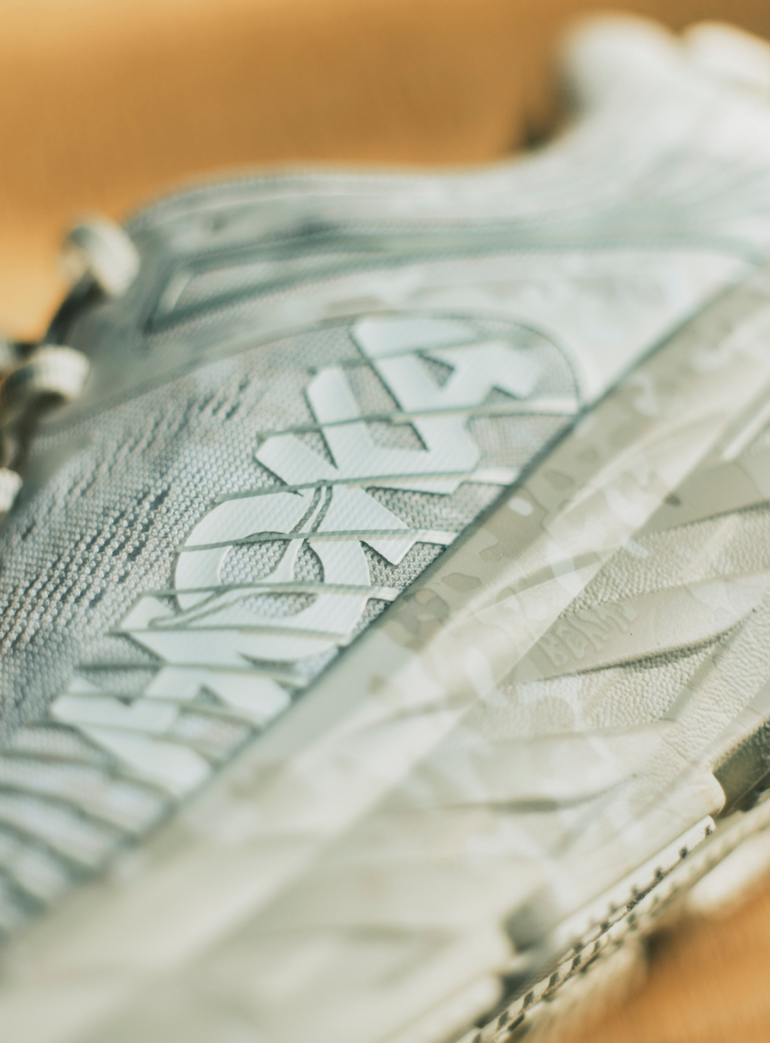 Close-up of a gray Hoka U BONDI 7 BEAMS sneaker, featuring a textured engineered mesh upper, visible branding, and part of a translucent, patterned midsole. The background is softly blurred.