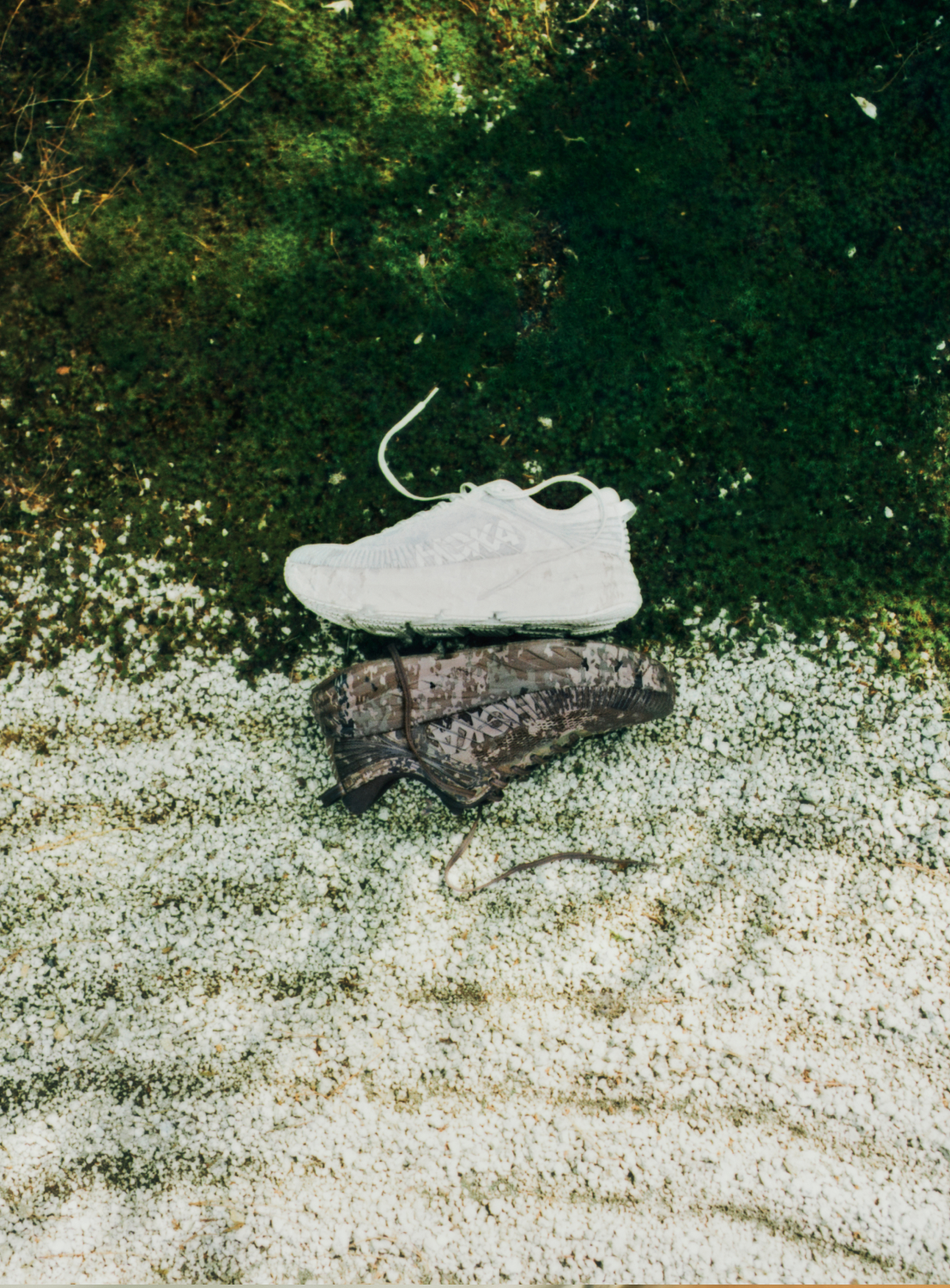 The Hoka U BONDI 7 BEAMS sneakers—one featuring a white engineered mesh upper and the other showcasing a dark, patterned design—are displayed side by side on a ground divided between green moss and white gravel, emphasizing their lightweight cushioning and unique style.