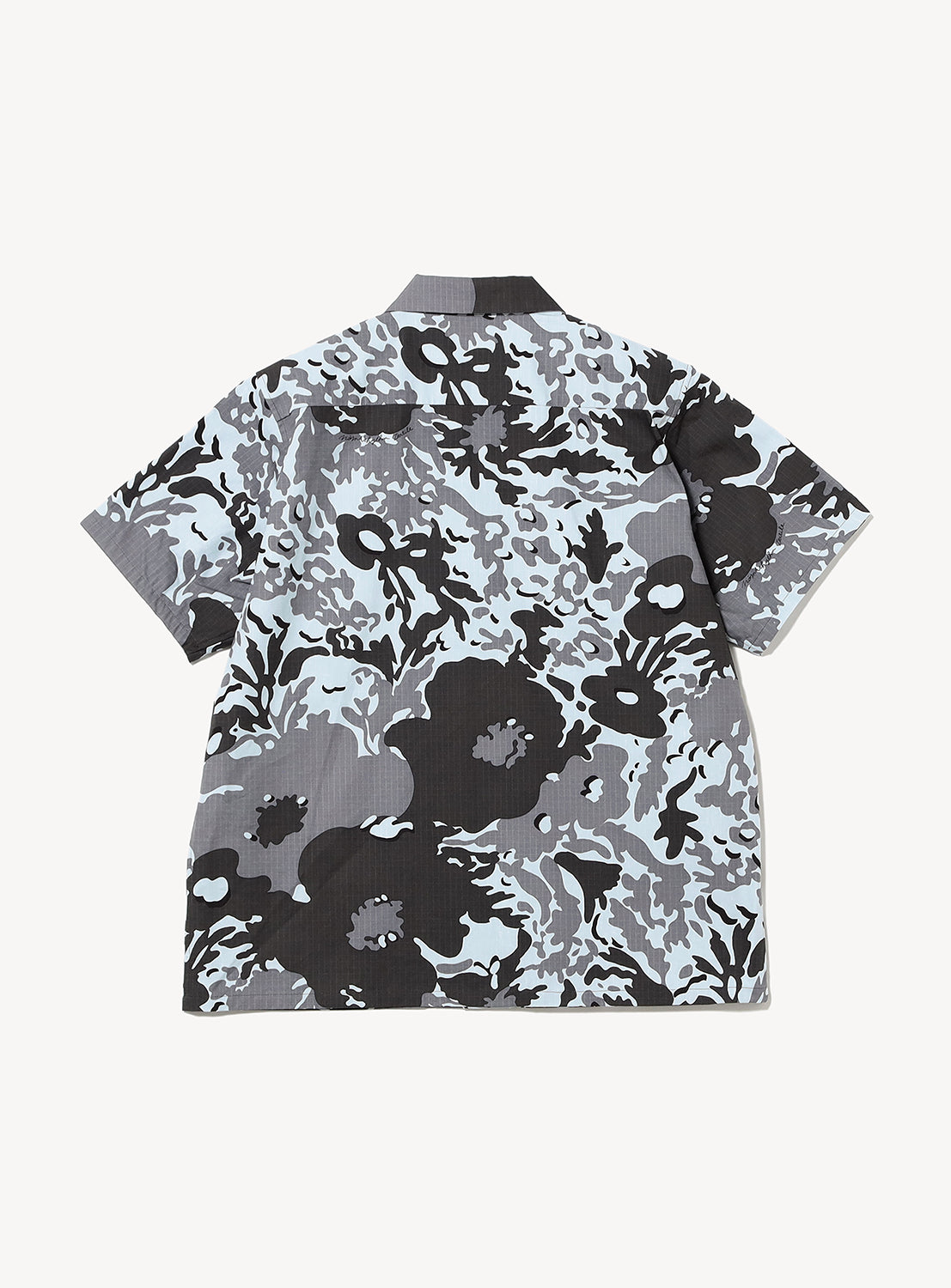 The Camo Flower Open Shirt by NOMARHYTHM TEXTILE is a short-sleeve button-up made in Japan from lightweight cotton, showcasing a black, white, and gray floral camouflage pattern. The shirt is laid flat on a white background with the backside visible.