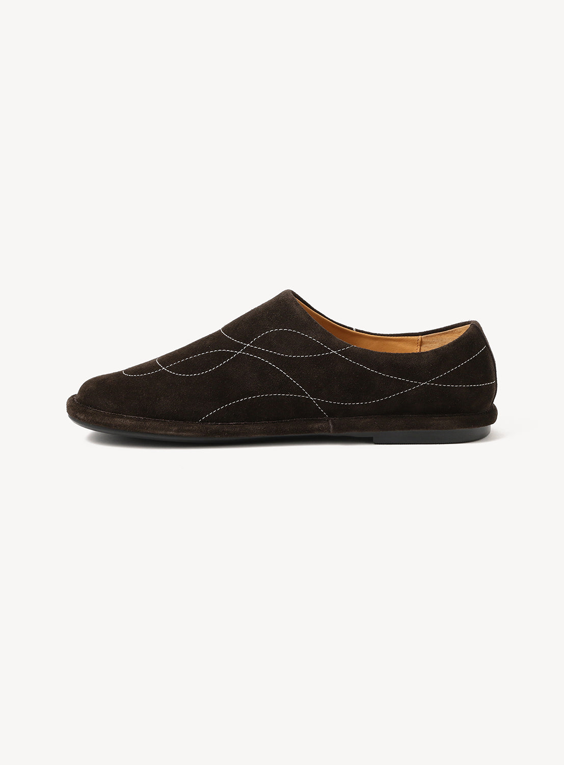 The OAO Sunlight Orb is a dark brown suede slip-on with white curved stitching, flat black sole, and tan lining. Its sculptural design is inspired by Isamu Noguchi and is shown in side profile against a plain white background.