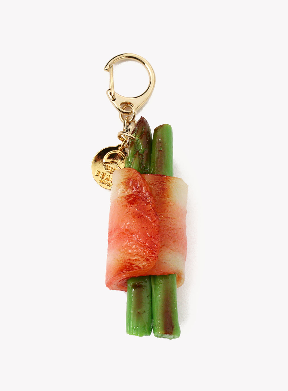 Gold-tone lobster clasp keychain charm shaped like two green asparagus spears wrapped in a thin slice of pink-and-white bacon. A small round gold tag hangs from the clasp. The charm is photographed against a plain light gray background.