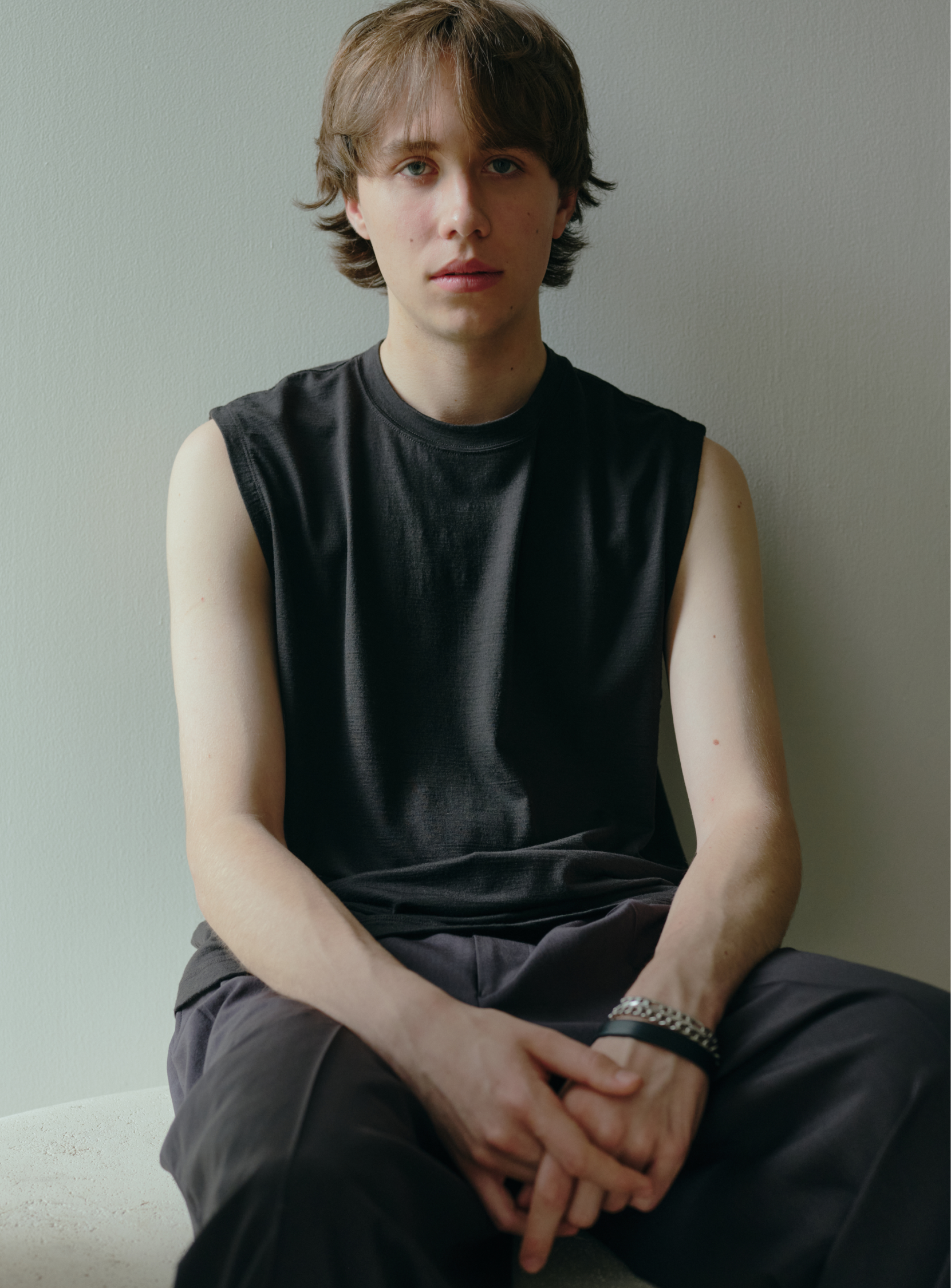 A young person with light skin and shaggy brown hair sits against a plain light wall, wearing BEAMS' the A - Washable Merino Wool Sleeveless Top and black pants, hands loosely clasped in their lap, with a calm expression.