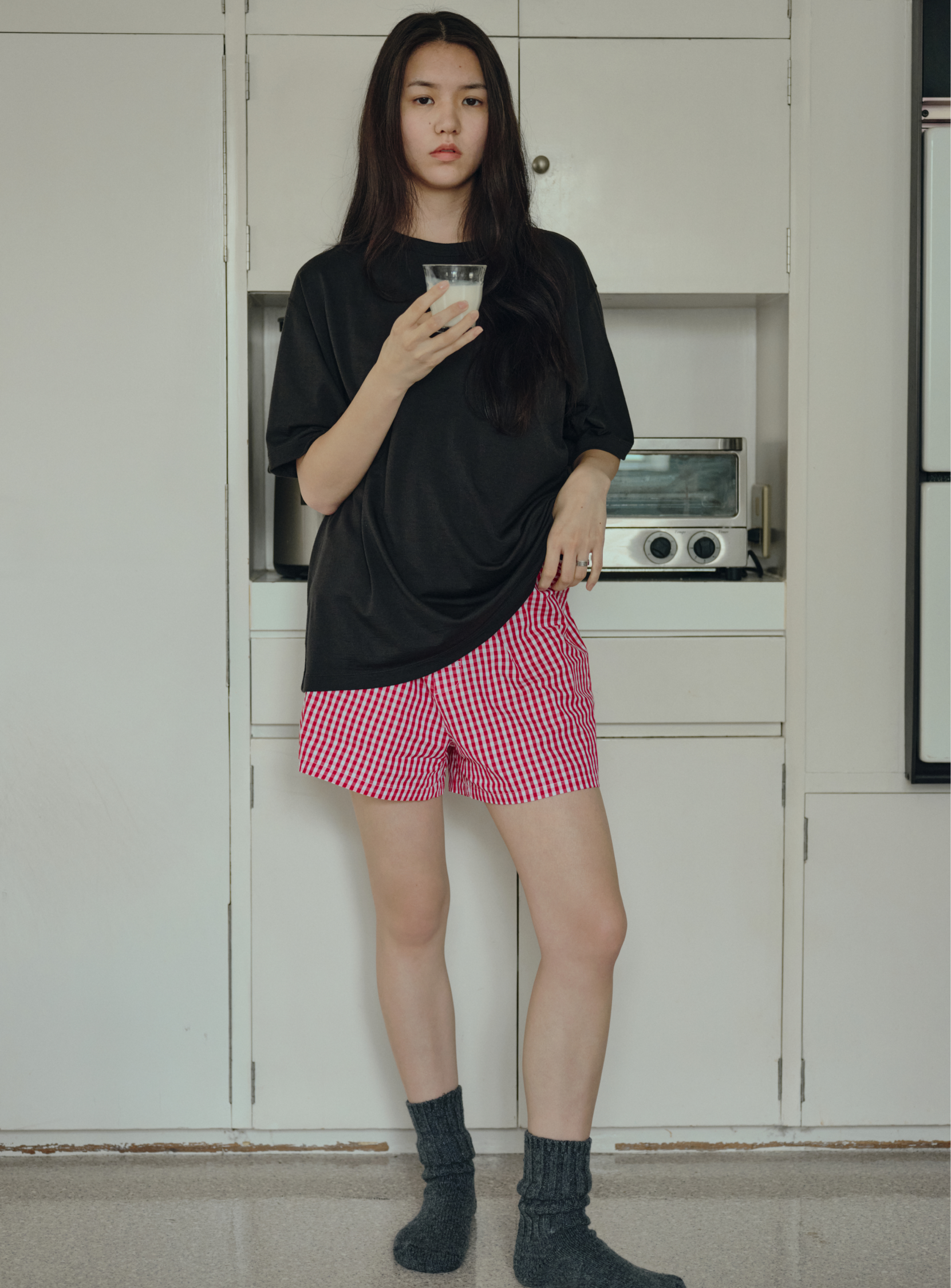 A young woman stands indoors holding a glass of milk. She wears BEAMS' the A - Washable Merino Wool T-Shirt in black, red checkered shorts, and gray socks. White cabinets and a toaster oven are visible behind her.