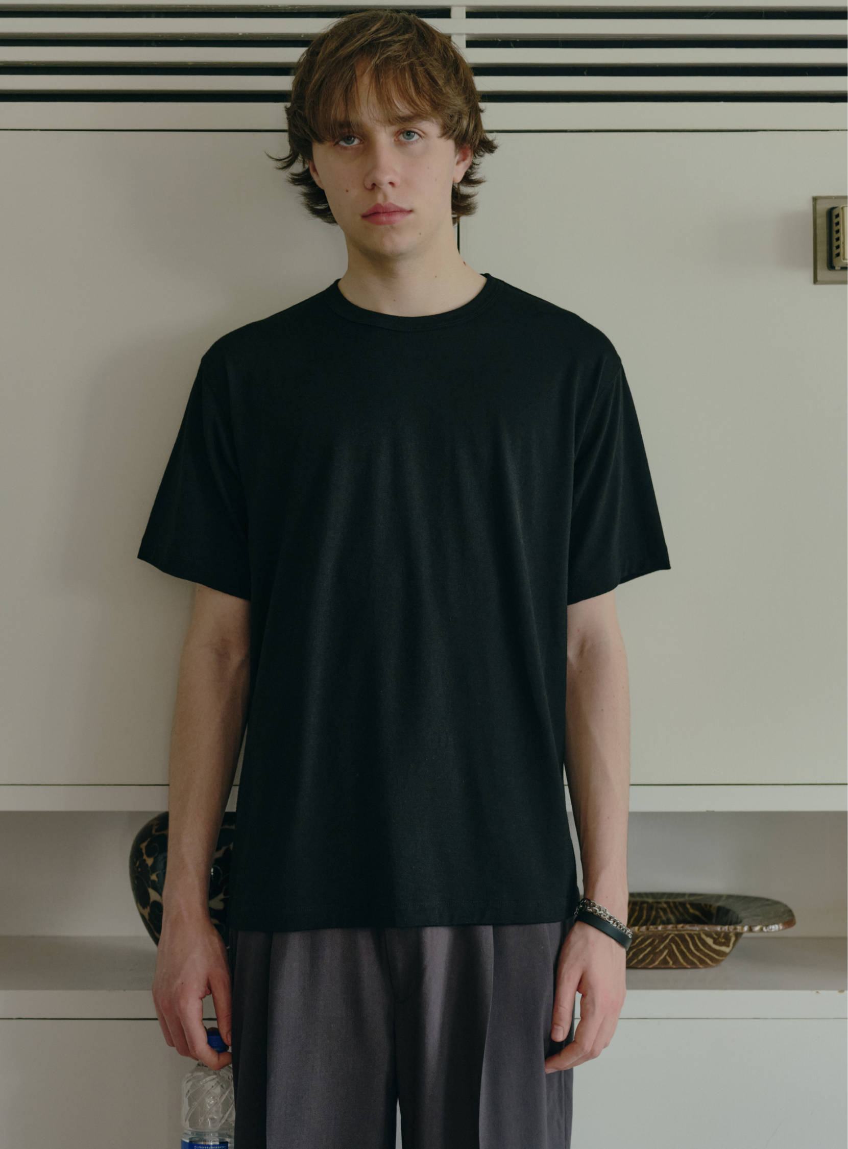 A young person with light brown hair stands indoors against a white wall, wearing BEAMS’ the A - Washable Merino Wool T-Shirt in black and gray pants, looking directly at the camera with a neutral expression.
