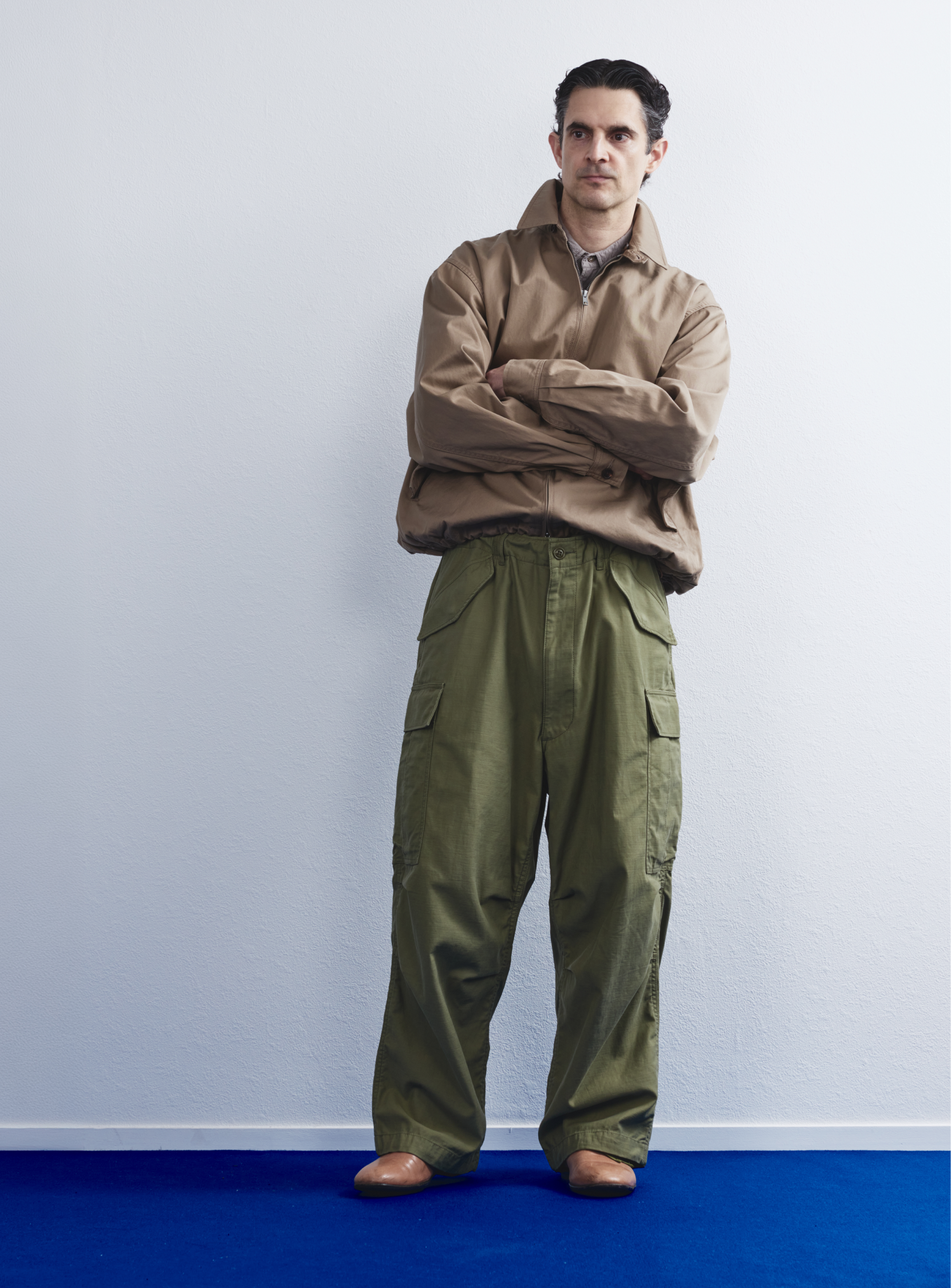A man stands with arms crossed, wearing a tan jacket, BEAMS the A - Big 6 Pockets Cargo Pants in olive green, and light brown shoes, set against a plain white wall and blue floor.