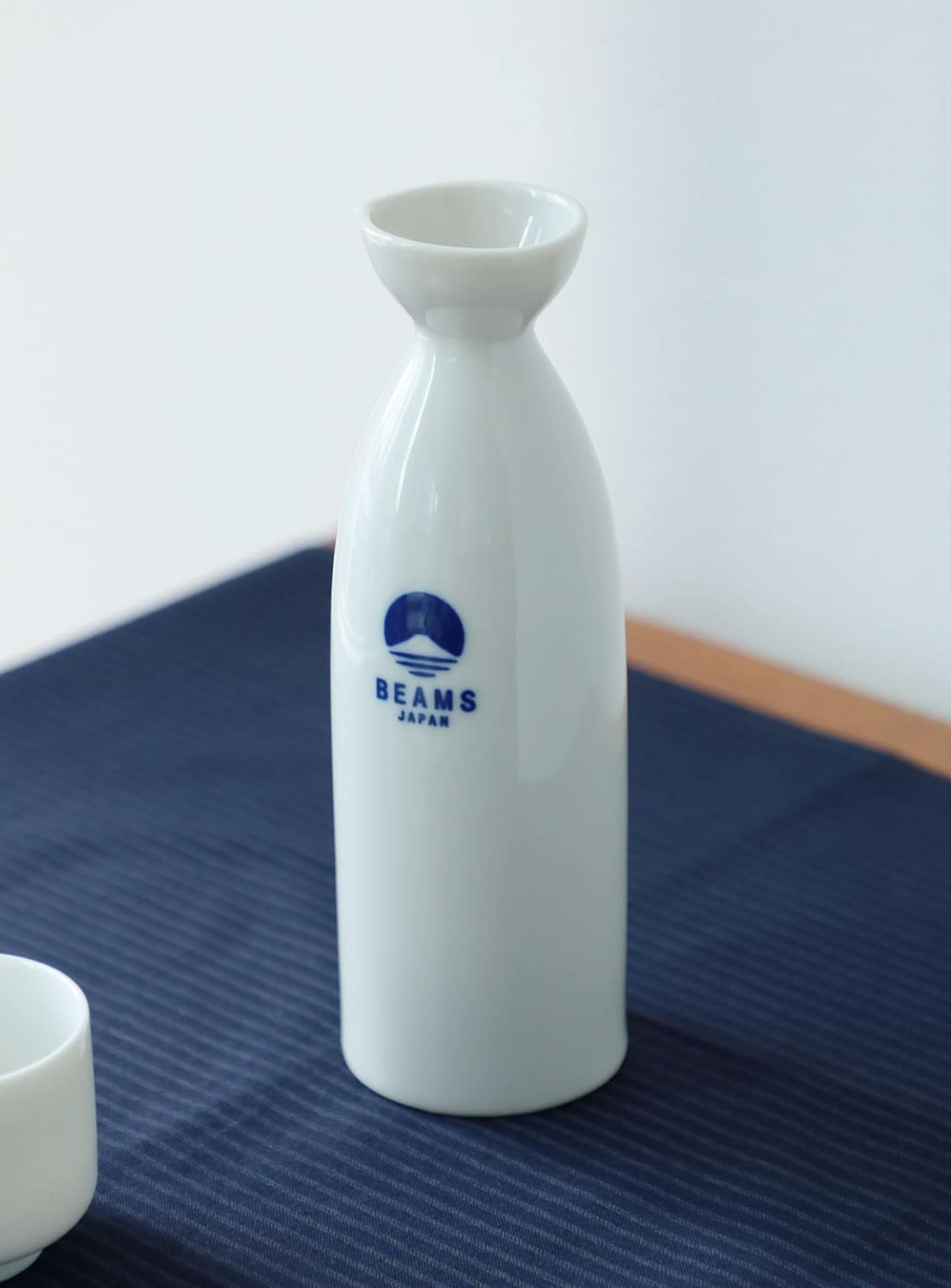 A BEAMS JAPAN Sake Bottle 360ML, crafted from white ceramic and adorned with a blue BEAMS JAPAN logo, rests on a dark blue cloth beside a small white cup—an elegant showcase of Japanese sake ware.