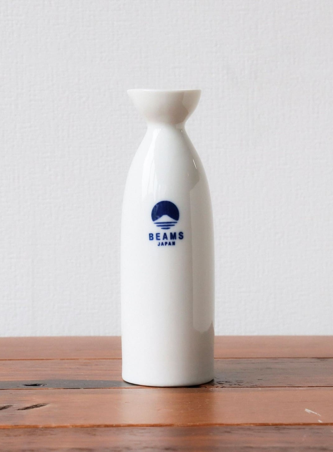 The BEAMS JAPAN Sake Bottle 360ML, crafted from white ceramic and featuring a minimalist tokkuri design, rests on a wooden surface set against a plain white background.