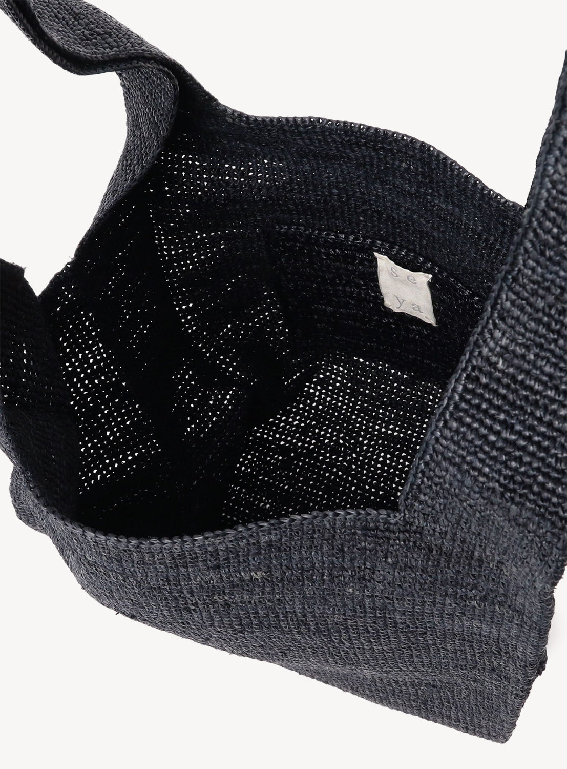 A close-up of the seya. Raffia Shopper Bag reveals its black handwoven raffia, fabric label on the lining, wide handles, and textured open weave—highlighting its unique craftsmanship.
