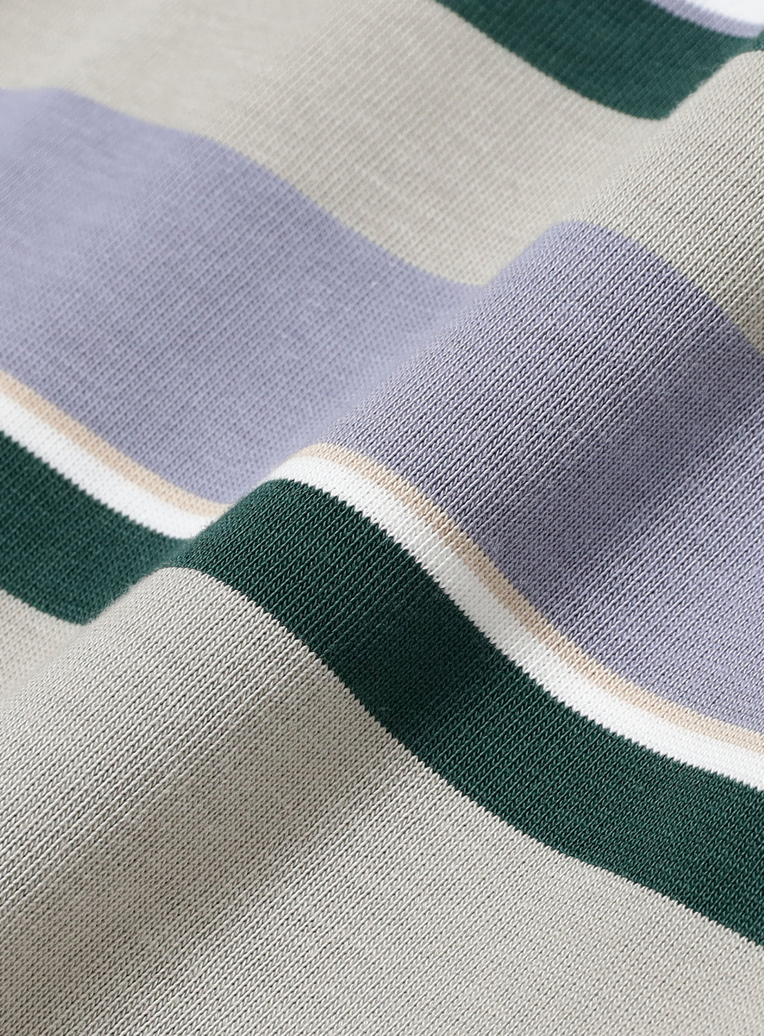 Close-up of the Multi-Border T-Shirt by BEAMS, showcasing its horizontal border stripes in gray, lavender, green, white, and beige. The 100% cotton fabric appears soft and slightly textured—typical of a premium crewneck T-shirt—with gentle folds that create subtle shadows and highlights across the surface.