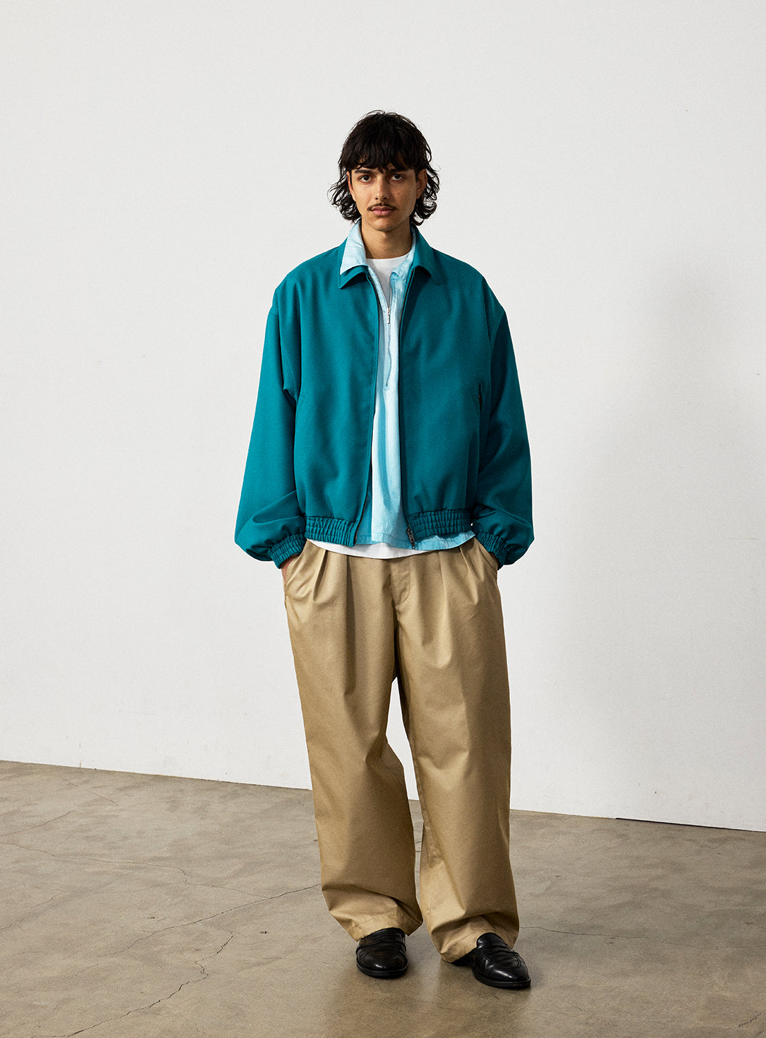 A person wearing a teal zip-up jacket over a light blue chambray shirt and tan pleated wide-leg BEAMS Chambray Chino Pants.